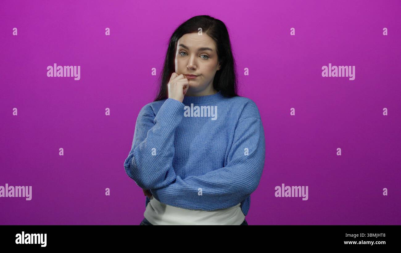 Woman standing against a vibrant pink background, wearing a blue sweater, with brunette hair, expressing contemplation with crossed arms and thoughtfu Stock Photo