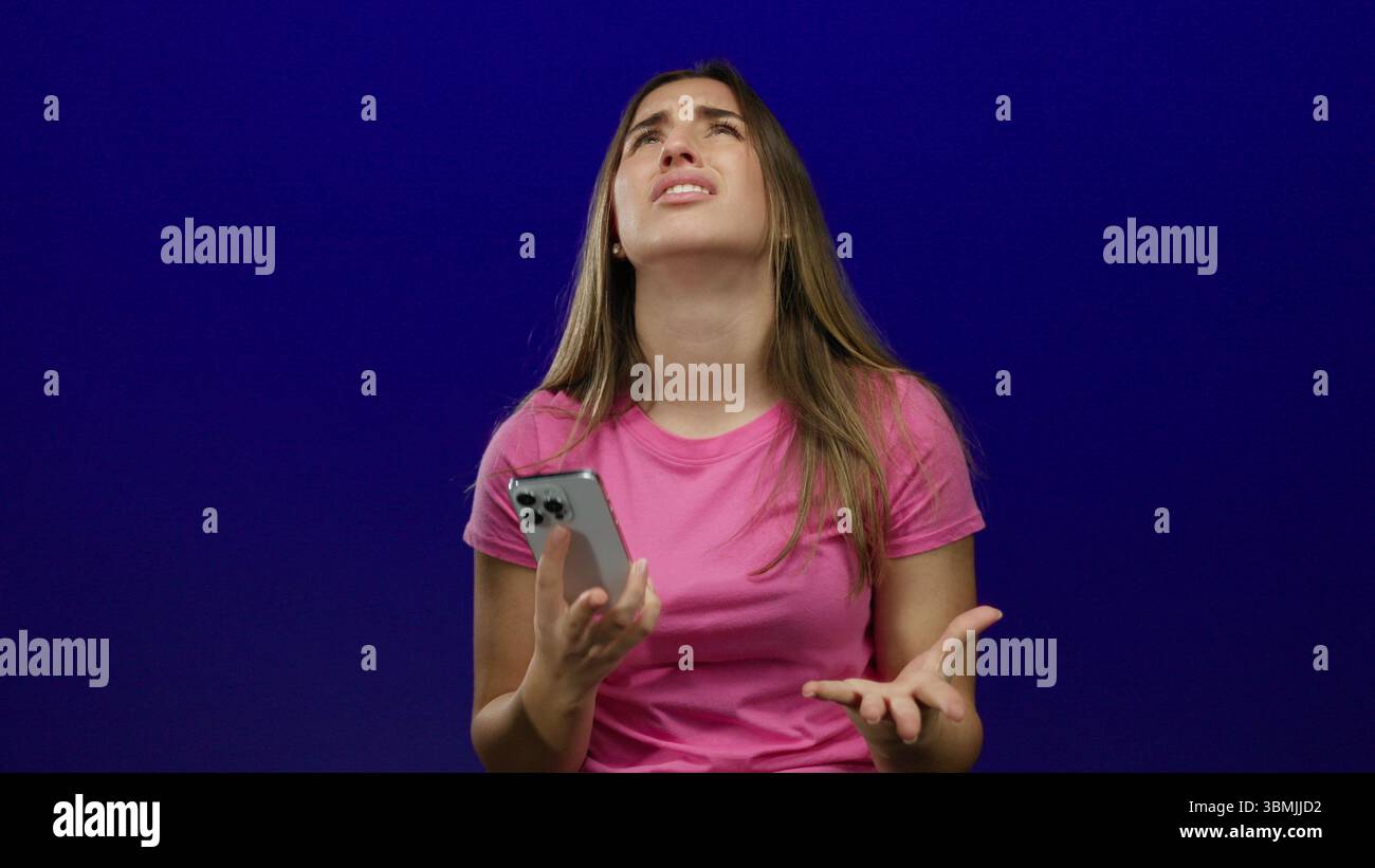 Woman expressing frustration while holding smartphone against blue background, wearing pink shirt, showcasing emotions of annoyance and confusion in a Stock Photo
