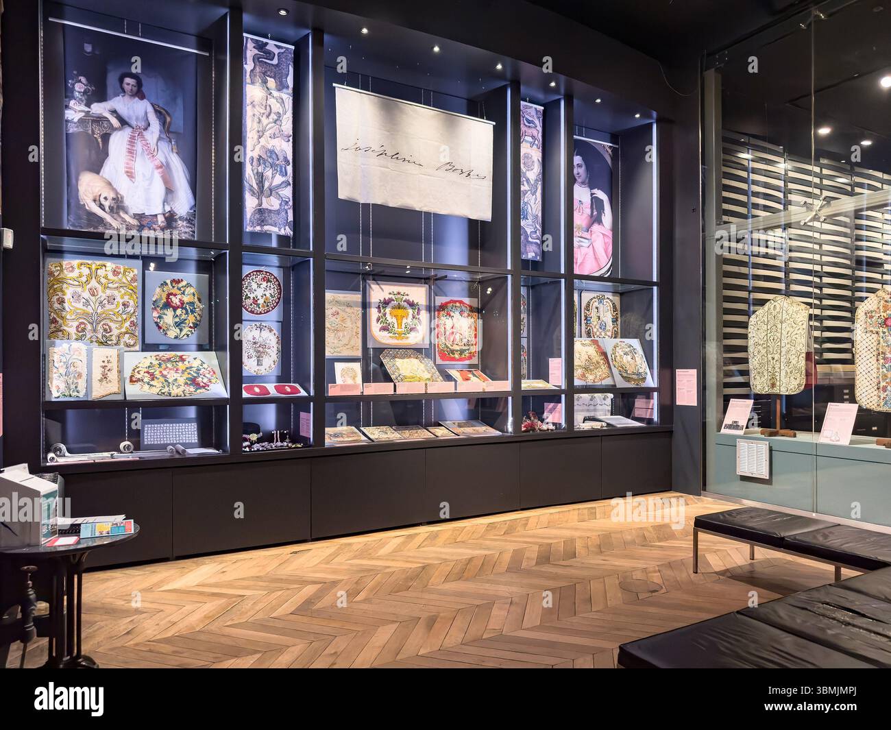 Exhibition space displaying ornate textiles and artworks in a well-lit glass case with wooden floor and modern decor - Smartphone Captured Stock Image