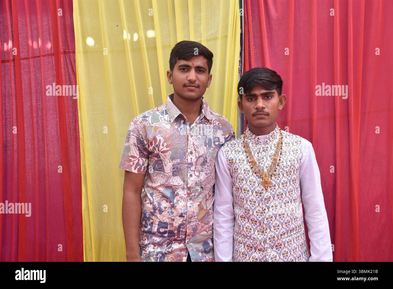 Two Young Indian Men Posing Together at Wedding Function with Colorful Drapes in Background Stock Photo