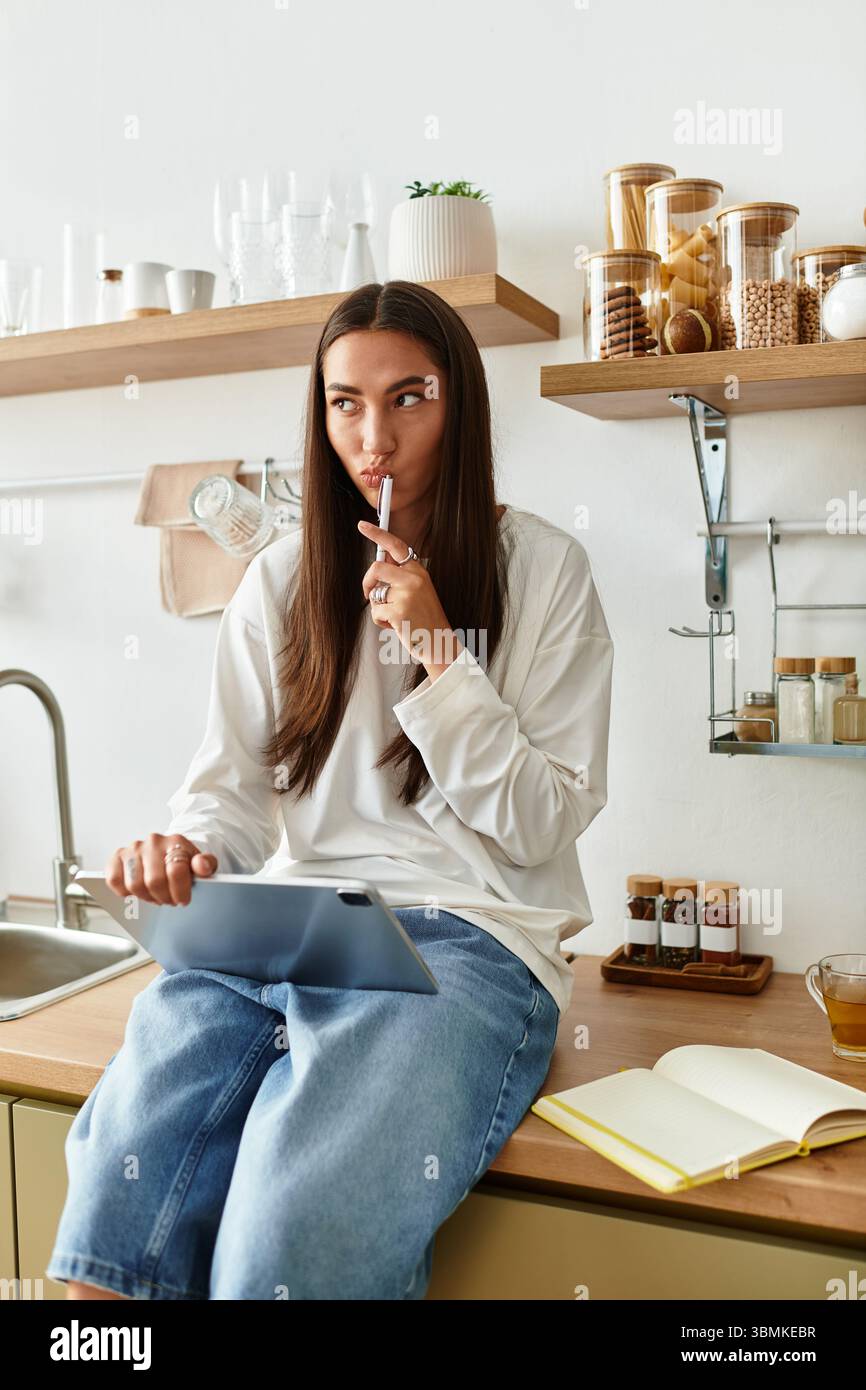A young woman sits thoughtfully in her homely kitchen, engaged with her tablet and pen. Stock Photo