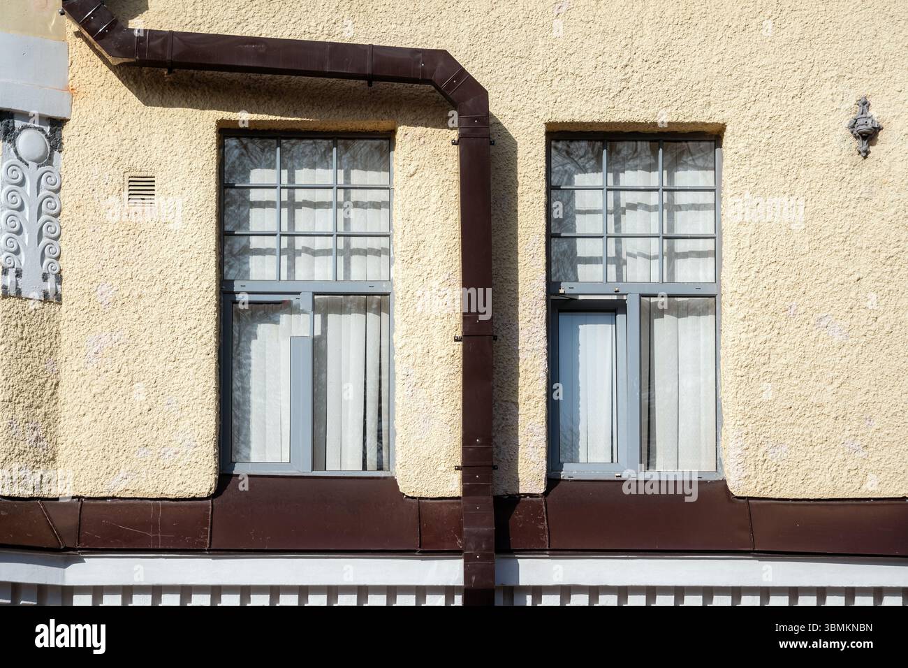 Two rectangular windows on a beige wall with a brown rectangular drainpipe. From the Windows of the World series. Stock Photo