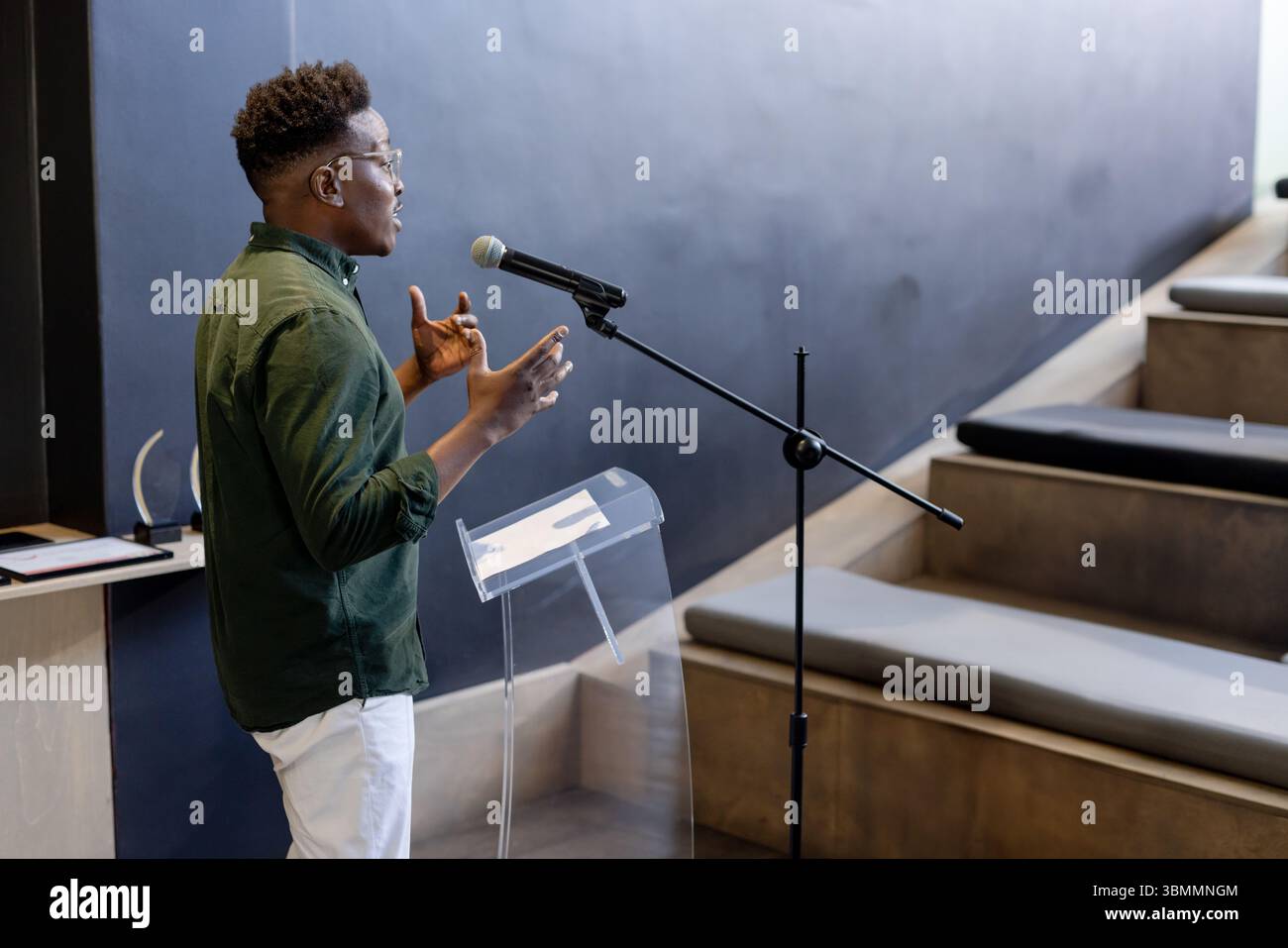 Male speaker gesturing at acrylic lectern with microphone, paper near award shelf at lecture hall Stock Photo