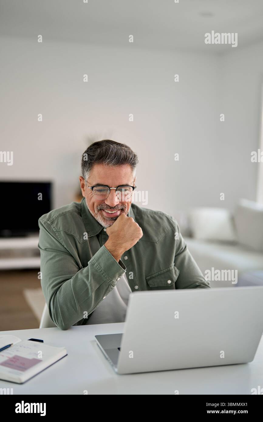 Happy middle aged man using laptop computer writing notes sitting at home table. Stock Photo