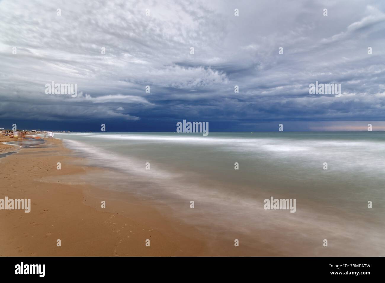 A long exposure captures the motion of the waves as they meet the shore under a dramatic, cloudy sky at the beach. Cut-off low Stock Photo