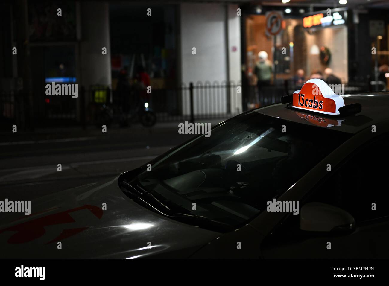 Illuminated orange 13 Cabs sign atop a taxi parked in a suburban shopping strip at night Stock Photo