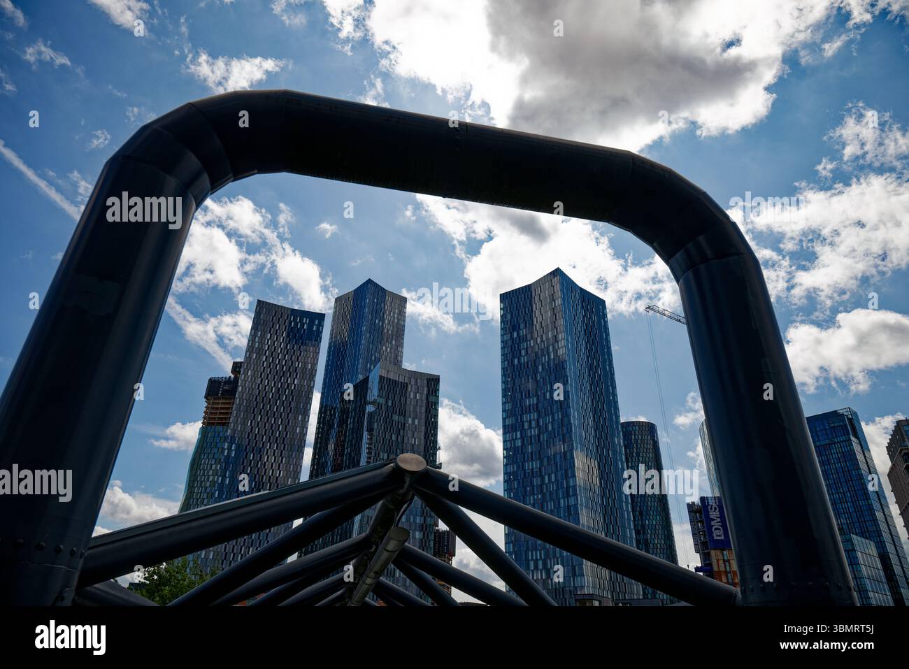 The skyscrapers around Deansgate Square rise into a sunny sky and are framed by the steel tubes of the footbridge to Deansgate Castlefield Station. Stock Photo