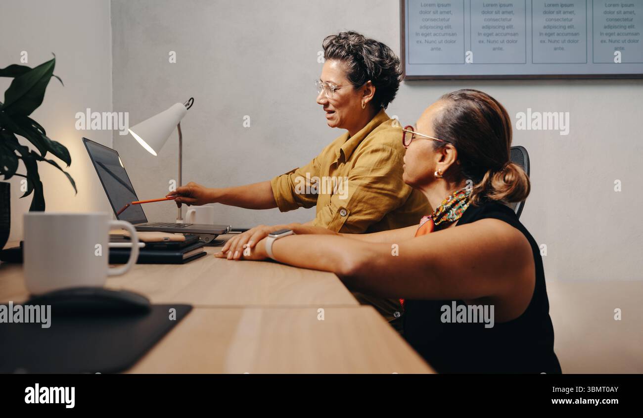 Two female professionals work together in an office on a project using a laptop. The teamwork highlights collaboration and productive interaction in a Stock Photo