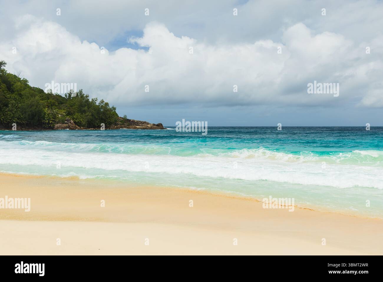 Mahe island, Seychelles. Peaceful tropical beach scene featuring golden sand, turquoise ocean waves, and lush green foliage under a cloudy sky, evokin Stock Photo