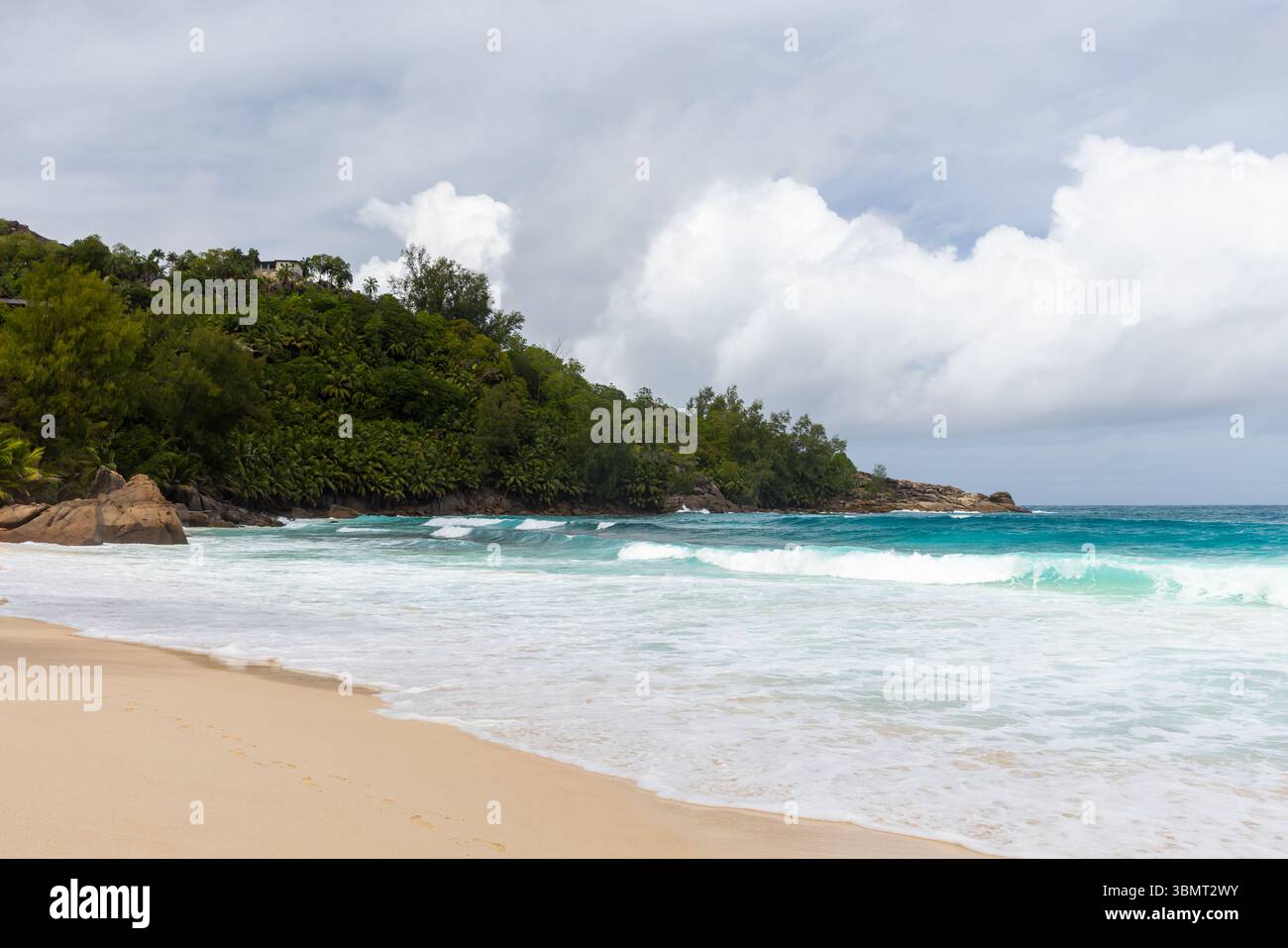 Mahe island, Seychelles. Tropical beach scene showcasing golden sand, turquoise waves, and green hillside vegetation under a partly cloudy sky, creati Stock Photo