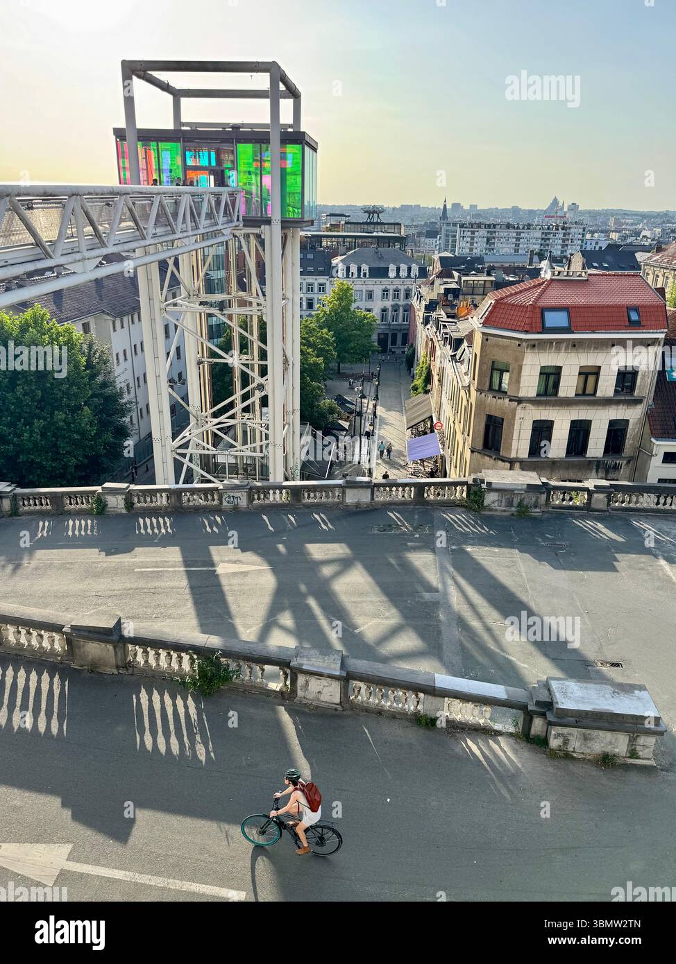 Cyclist riding below the panoramic glass elevator at Place Poelaert, offering a scenic view over Brussels, Belgium on a bright summer day. - Smartphone Captured Stock Image