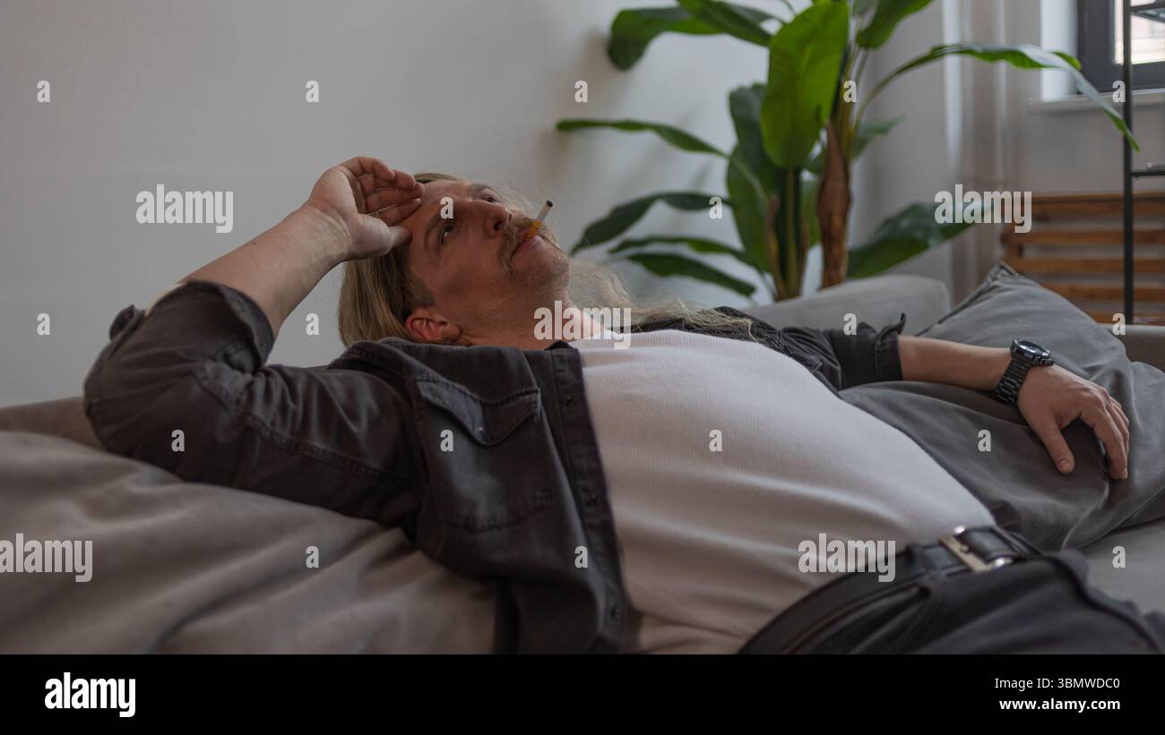 An unkempt man in deep depression sprawled on the couch, a limp pose speaks  of a breakdown and lack of meaning in life, a lit cigarette in his mouth  Stock Photo -