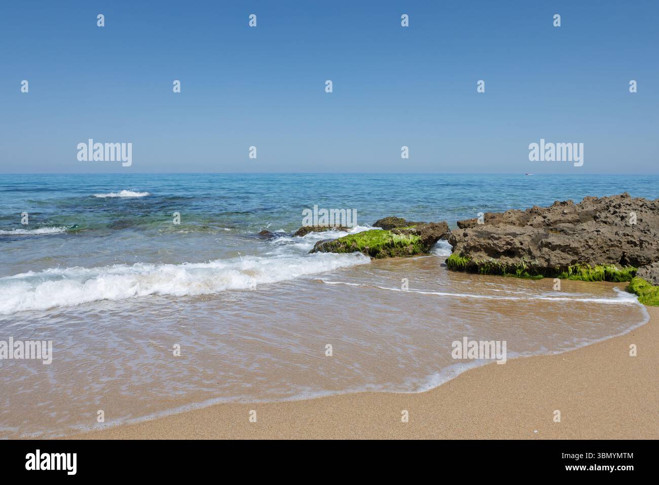 Waves hitting algae-covered rocks on a sunny coastal beach with clear turquoise water Stock Photo
