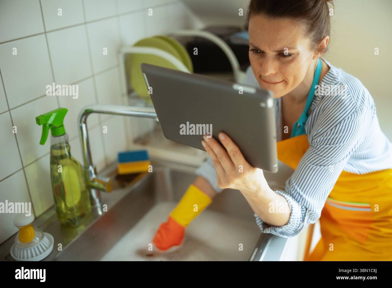 A woman in gloves and apron uses a tablet while facing a clogged kitchen sink, searching for help or instructions to resolve a drain issue. Stock Photo