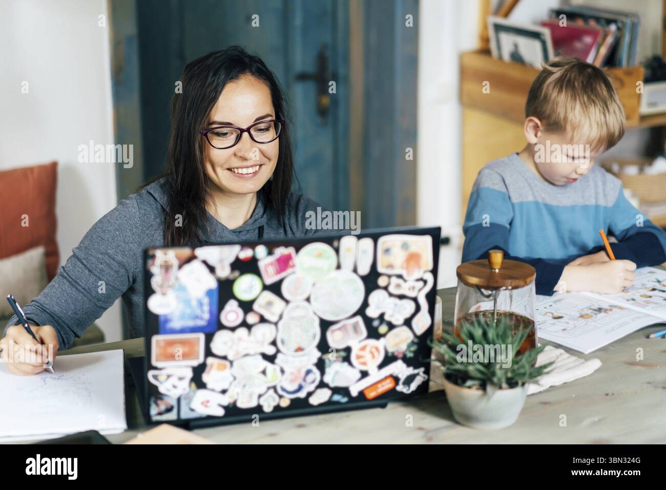 Work from home and family education. Mom and son are sitting at the desk. Business woman works in a laptop, a child writes in a textbook. Remote acces Stock Photo