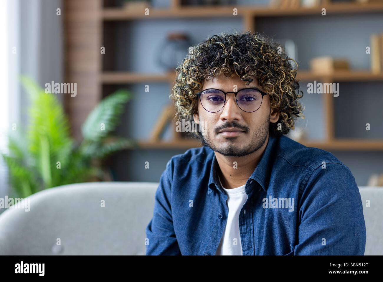 A young man with curly hair and glasses poses indoors, looking directly ...