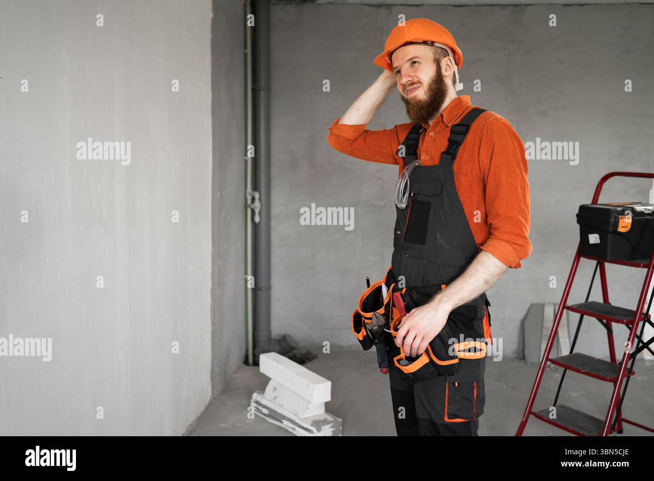 Bearded repairman or builder in protective helmet and overalls having doubts with confuse face expression scratching head in the room with building eq Stock Photo