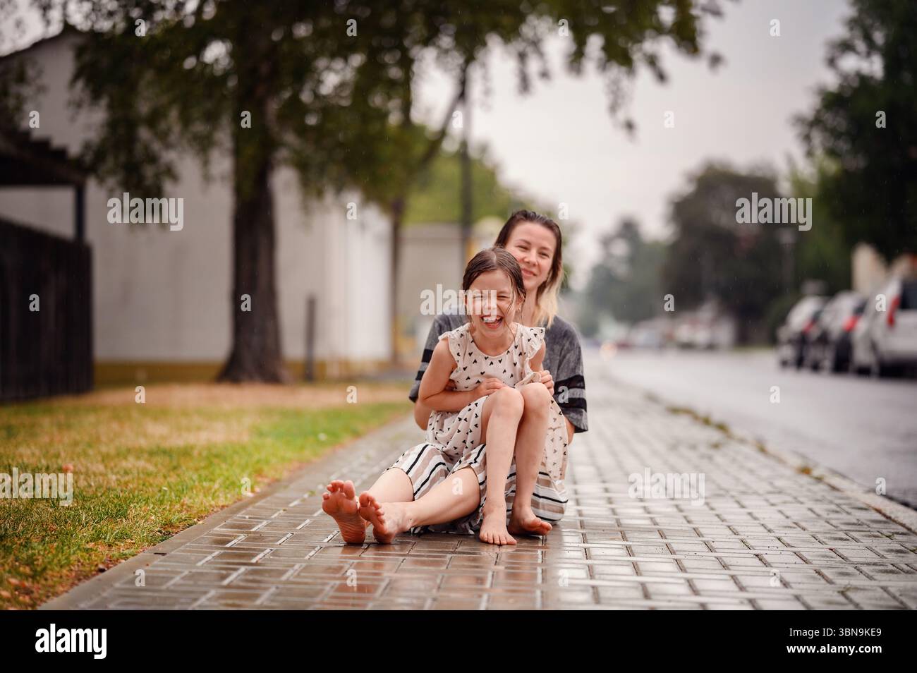 Smiling Mother And Daughter Sit Barefoot On A Wet Sidewalk Laughing smiling-mother-and-daughter-sit-barefoot-on-a-wet-sidewalk-laughing