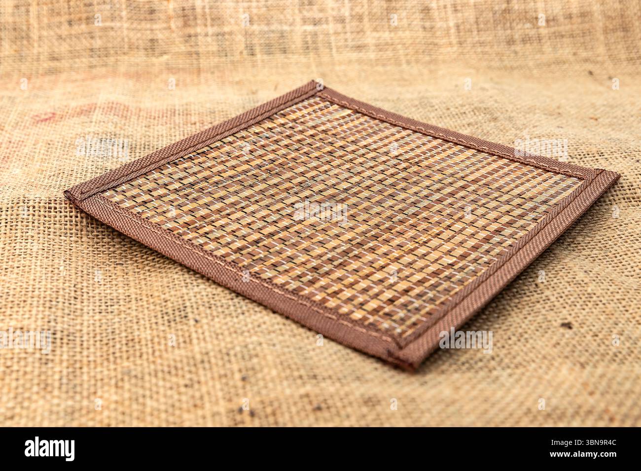 Square handmade woven placemat on burlap fabric, rustic natural background for kitchen, dining, table setting, eco decor and cozy home interior. Stock Photo