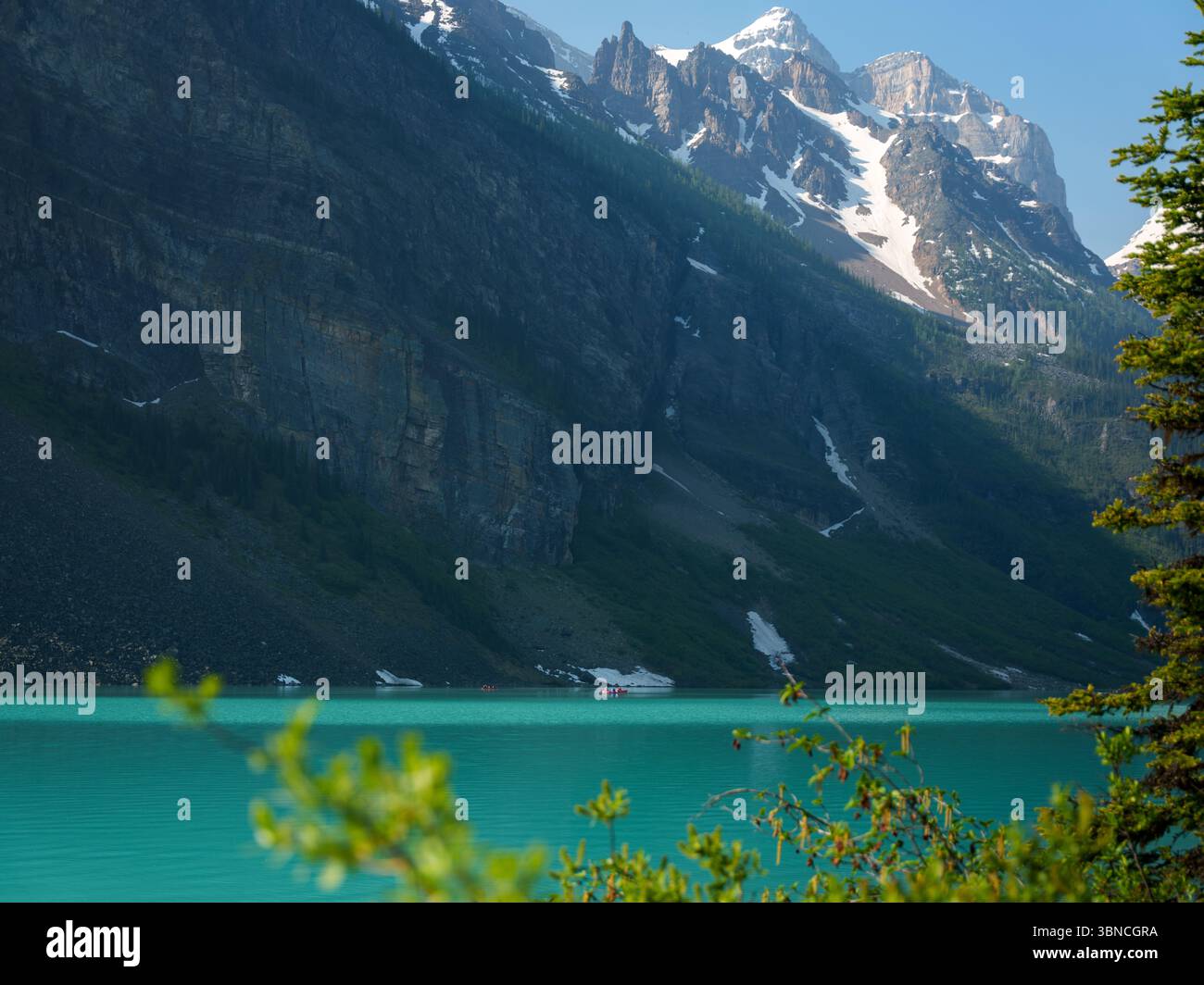 Steep rocky cliffs towering over the turquoise waters of Lake Louise, framed by green foliage in the foreground. A striking view from Banff National Stock Photo
