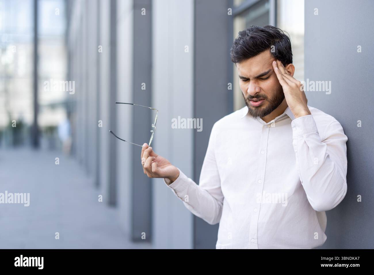 A man experiencing a headache, holding glasses, and leaning against a wall outside of a building. Stock Photo