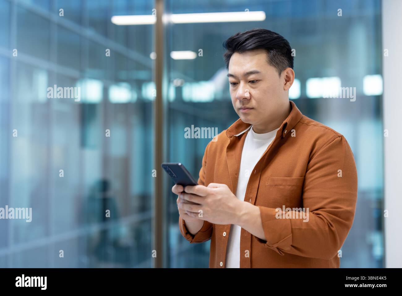 An Asian man uses his smartphone inside a modern office. He is dressed casually and seems focused on his phone. Stock Photo