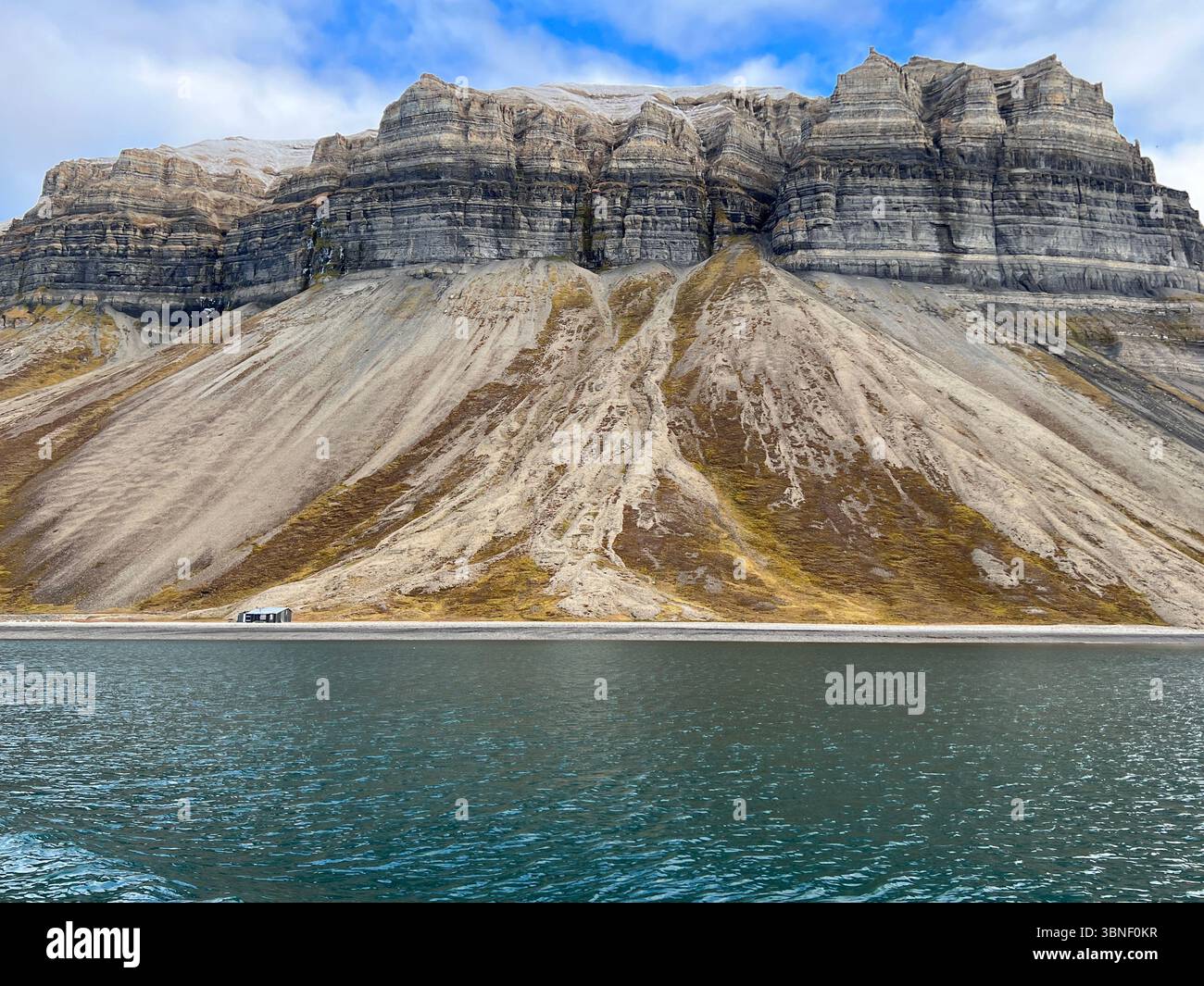 A small cabin is situated beside the ocean with a stunning mountain in Nordre Isfjorden, Spitsbergen, Svalbard - Smartphone Captured Stock Image