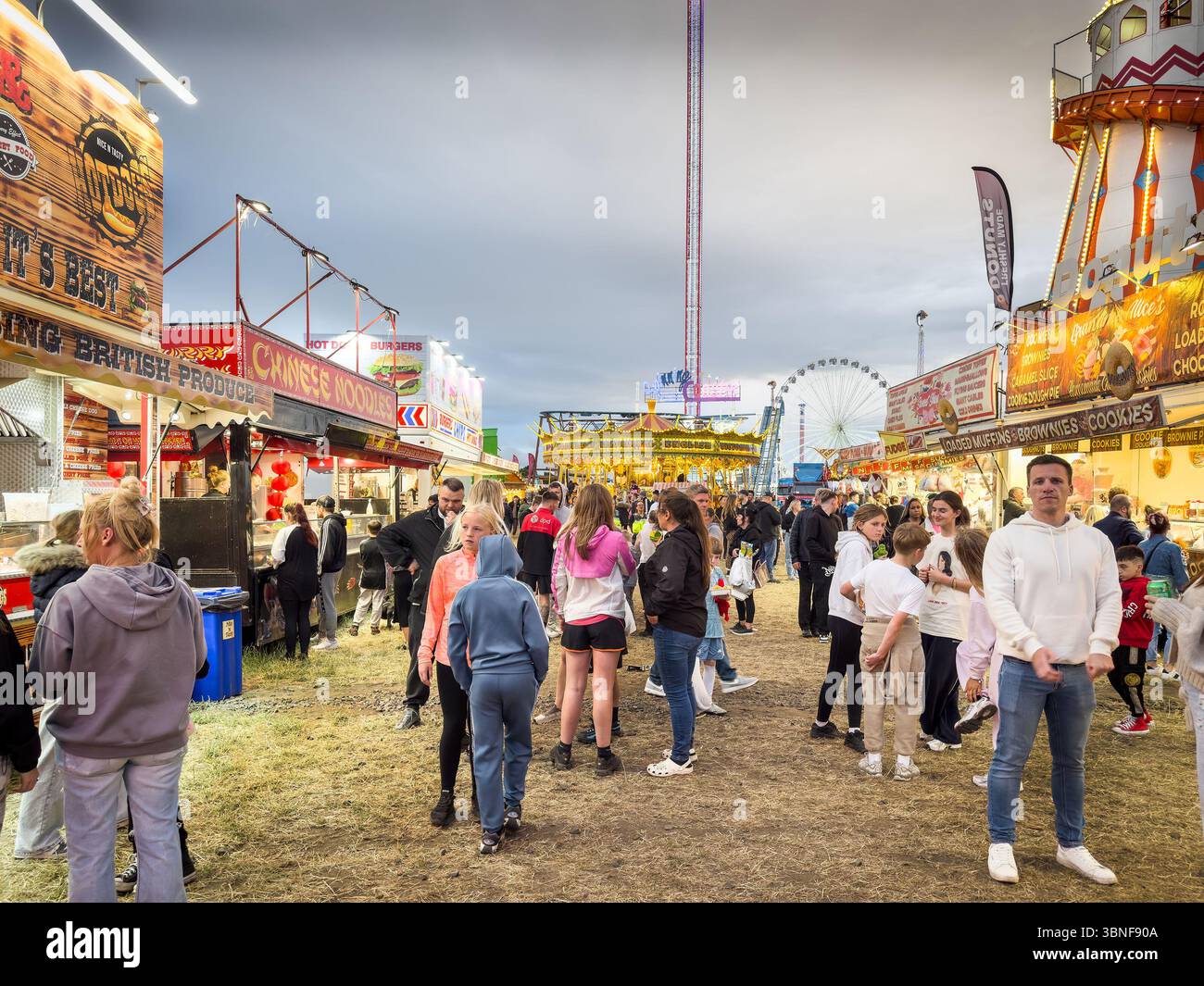 Crowd exploring food stalls and amusement rides at bustling evening fairground with ferris wheel & lively atmosphere The Hoppings town moor newcastle - Smartphone Captured Stock Image