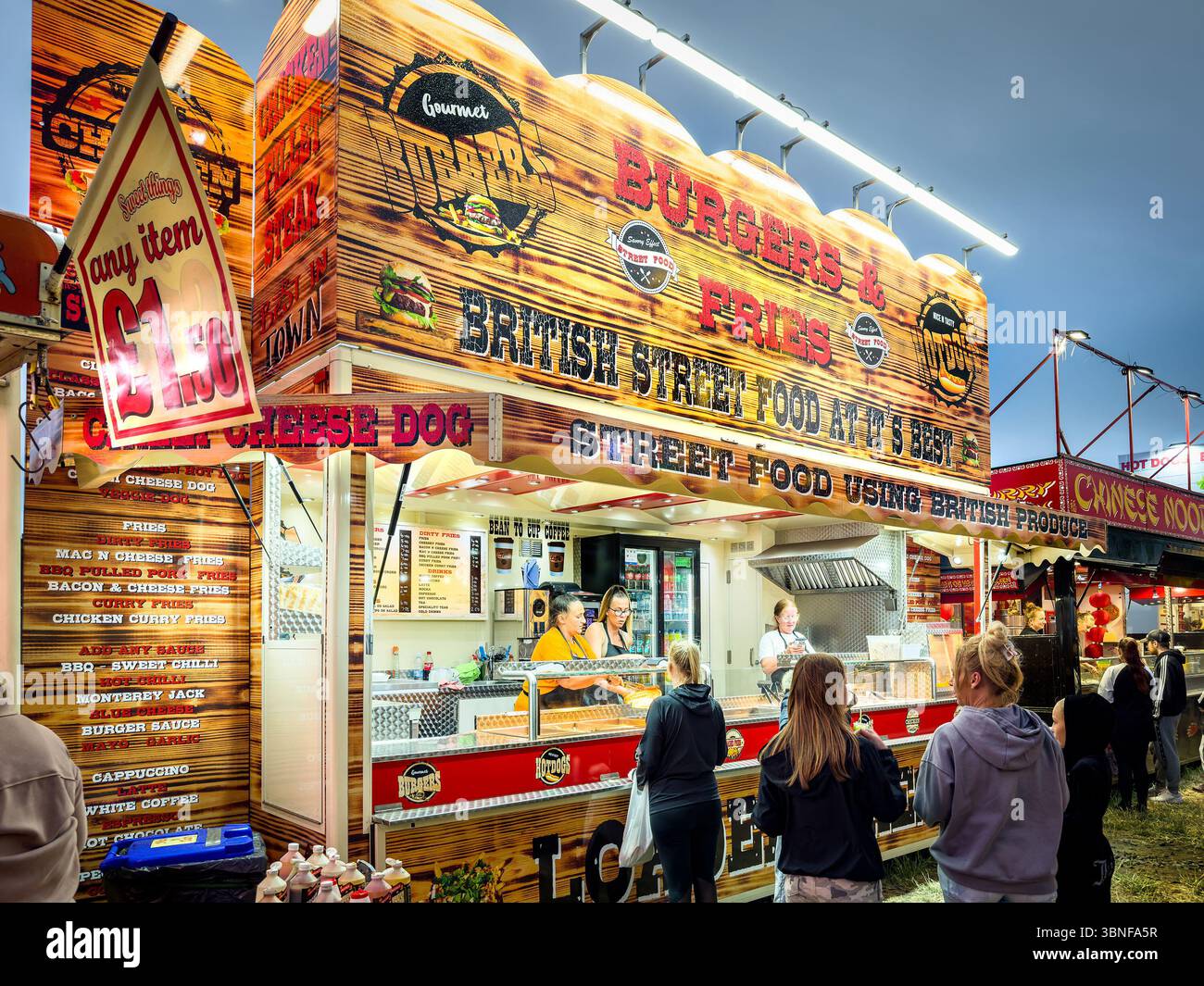 People standing under a brightly lit street food stall offering British cuisine at a local fair during the evening The Hoppings town moor newcastle - Smartphone Captured Stock Image
