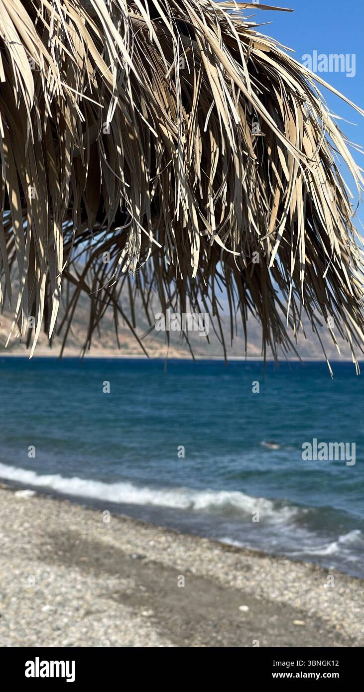 Close-up of a straw umbrella providing shade on a sunny beach day, with gentle waves rolling onto the shore in the background. The image captures a pe Stock Photo
