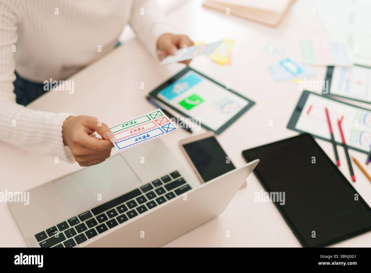 Asian adult woman hands holding user interface card for mobile app testing on desk with laptop tablet and smartphone. Concept for digital design proce Stock Photo