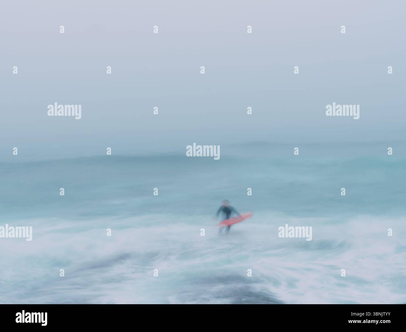 A lone blurred surfer figure amongst the waves in a blue white seascape with movement Stock Photo
