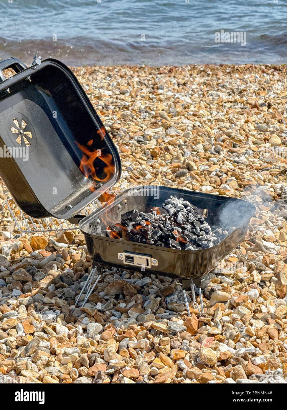Unattended barbecue with flames from hot coals at the water's edge of a shale beach - Smartphone Captured Stock Image