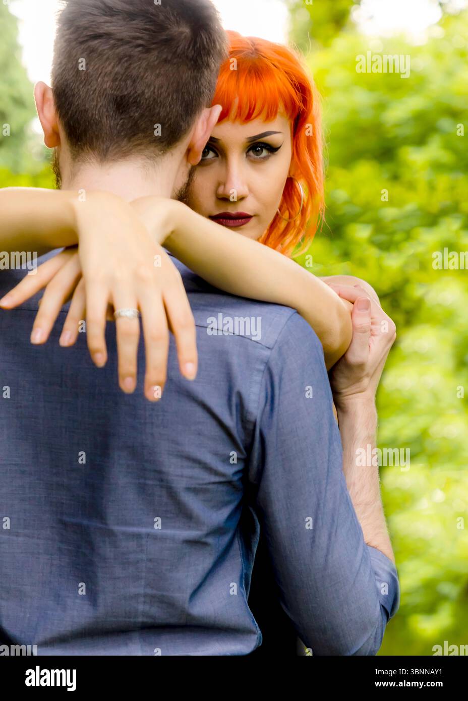 A woman with bright orange hair embraces a man from behind, her arms ...