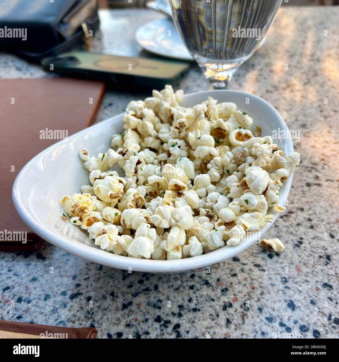 A modern white, boat-shaped bowl holds a generous serving of freshly popped, seasoned popcorn, lightly sprinkled with finely chopped green herbs, like - Smartphone Captured Stock Image