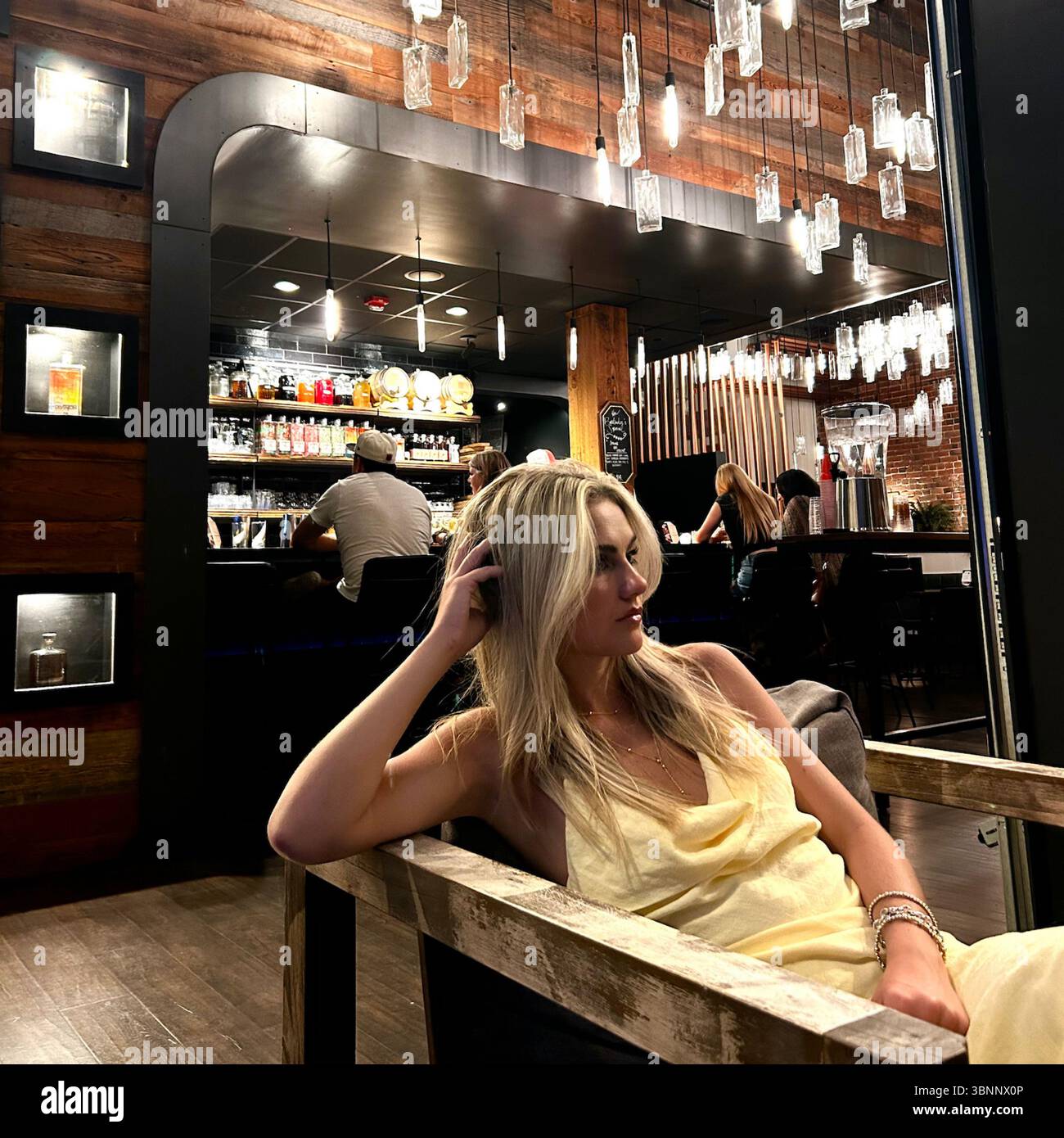 A woman in a soft yellow dress sits casually in a wooden chair at a chic bar lounge with industrial-modern design elements. Overhead, unique pendant. - Smartphone Captured Stock Image