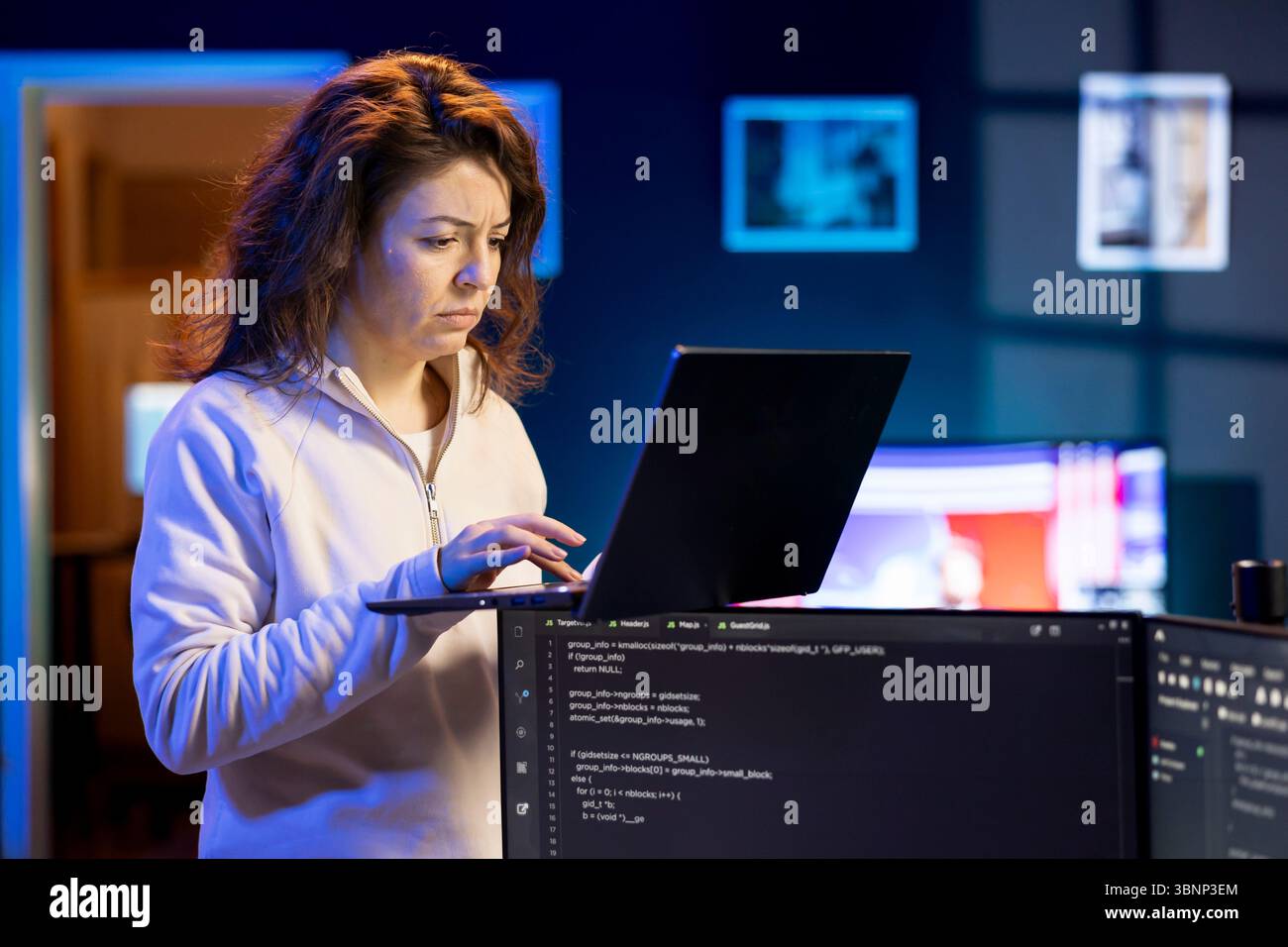 IT system administrator doing software debugging using laptop, checking glitches in lines of code displayed on desktop PC monitors. Woman standing in office at home fixing coding issues using notebook Stock Photo