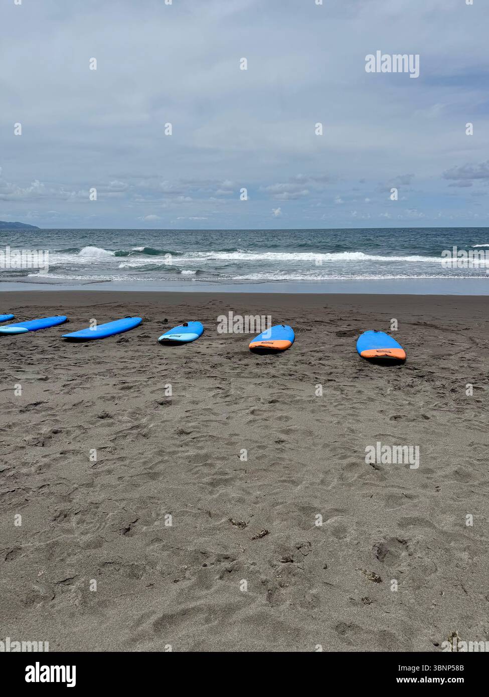 A row of surfboards lying on the volcanic sand beach of São Miguel Island, Azores, Portugal. Atlantic Ocean waves roll in under a partly cloudy sky. - Smartphone Captured Stock Image