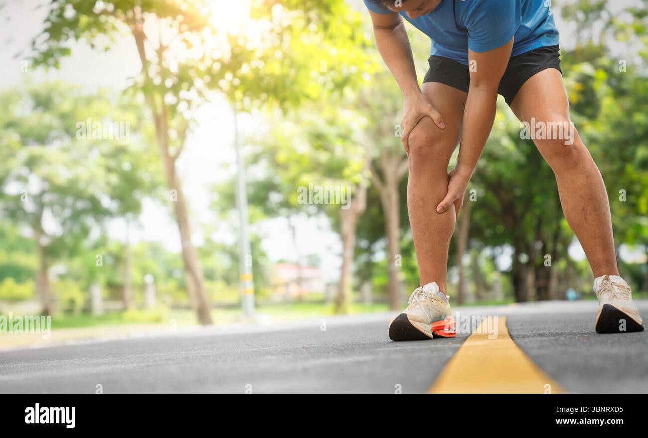 Athlete bending forward clutching calf muscle feeling discomfort while jogging on sunny outdoor road Stock Photo