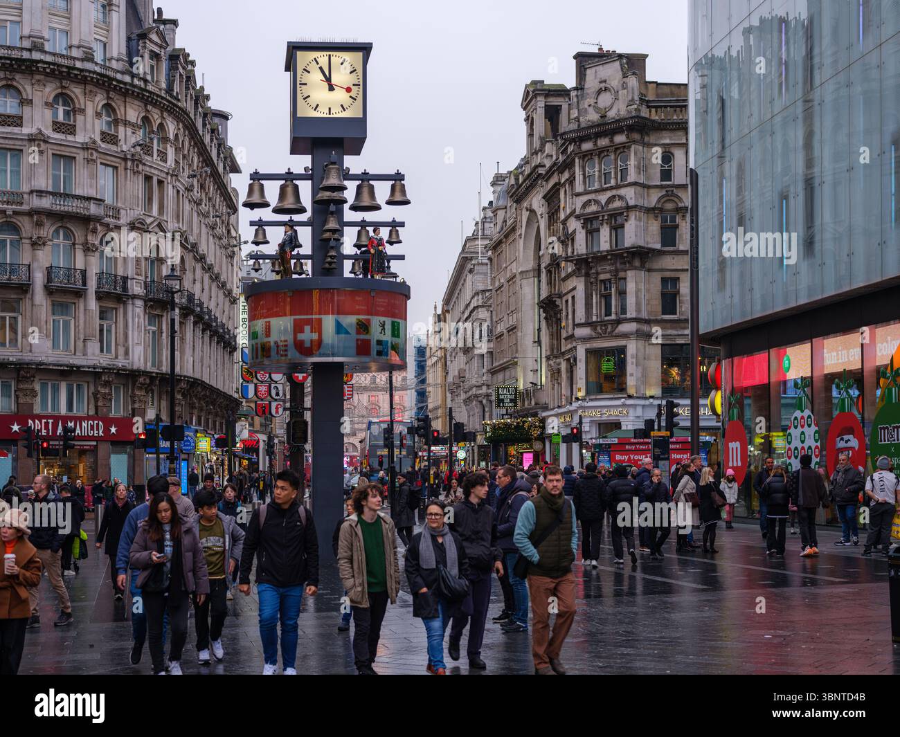 United Kingdom, London, 09 Jul 2025. Crowds walk past the Swiss Glockenspiel in Leicester Square on a rainy day, surrounded by historic architecture a Stock Photo