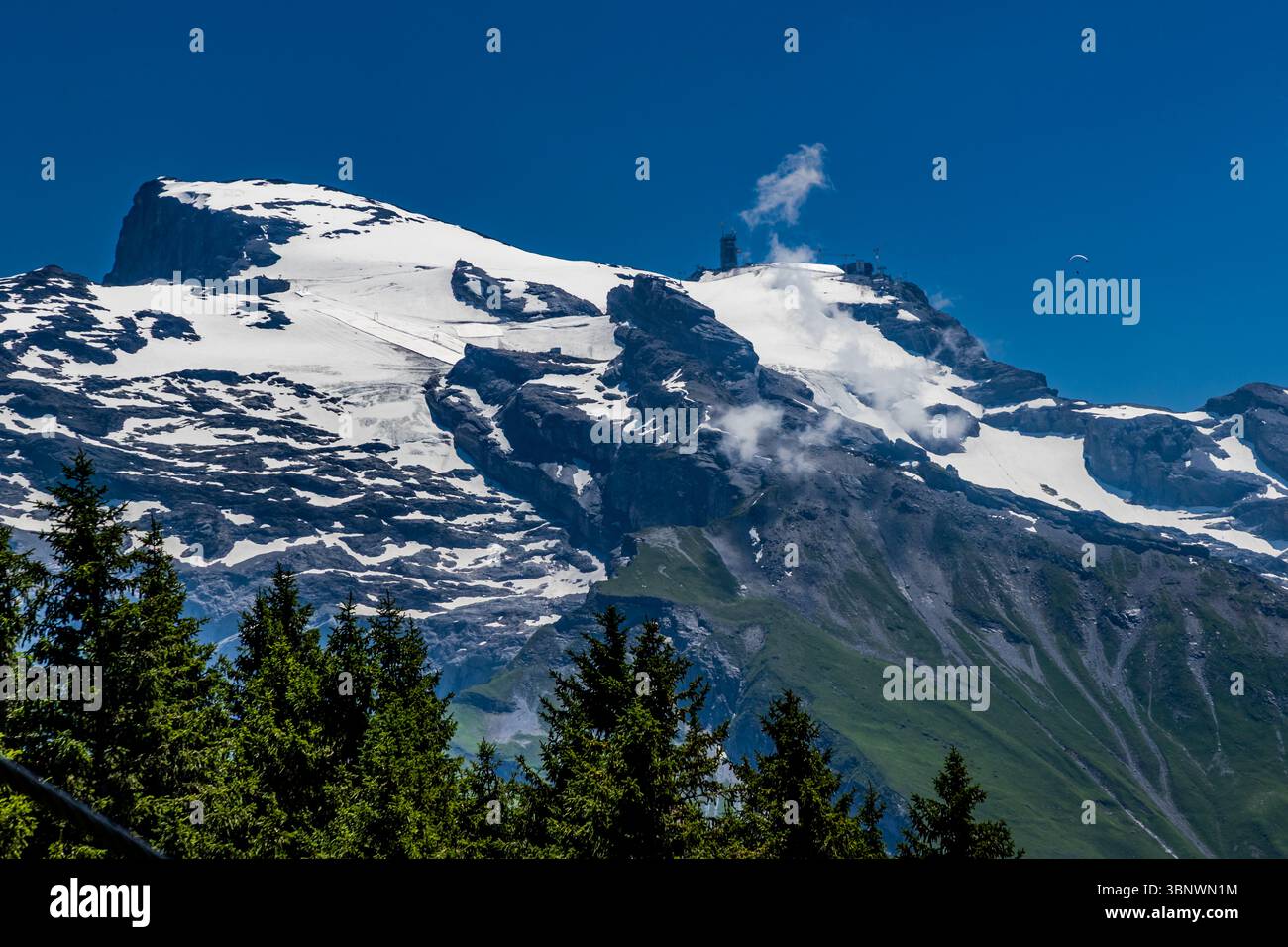 The Titlis is the heart of a large ski area with 82 kilometres of slopes. Attractions at the summit include the Titlis Cliff Walk – Europe's highest suspension bridge – and a glacier grotto. In 2025, however, the summit will be a huge construction site. The Titlis 3020 construction project involves the construction of a new mountain station at an altitude of 3,020 metres and a new cable car. The Titlis Tower is scheduled to open in May 2026. A paraglider soars over the impressive, snow-covered mountain landscape of the Titlis massif in Engelberg, Obwalden, Switzerland. The glacier and the moun Stock Photo