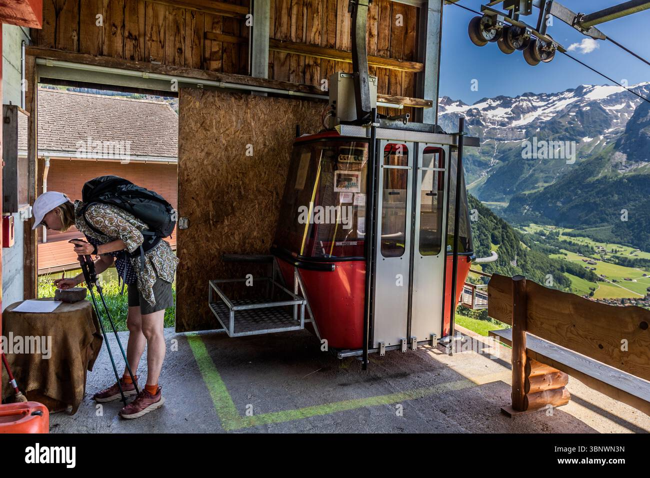 In the Engelberg Valley, a small lift system connecting remote farms with the valley is called a Bauernbahn, known in Swiss dialect as a Buiräbähnli. Hikers can use the lift with a valid lift ticket. One person with a rucksack and hiking poles signs in at a gondola lift station. The gondola lift is self-service. The station is located in Engelberg, Obwalden, Oberes Bord, with a view of the Swiss Alps Stock Photo