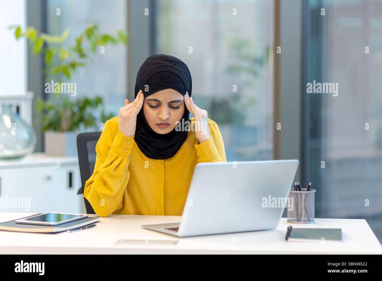 A woman in a hijab at her desk, appearing stressed while working on her ...