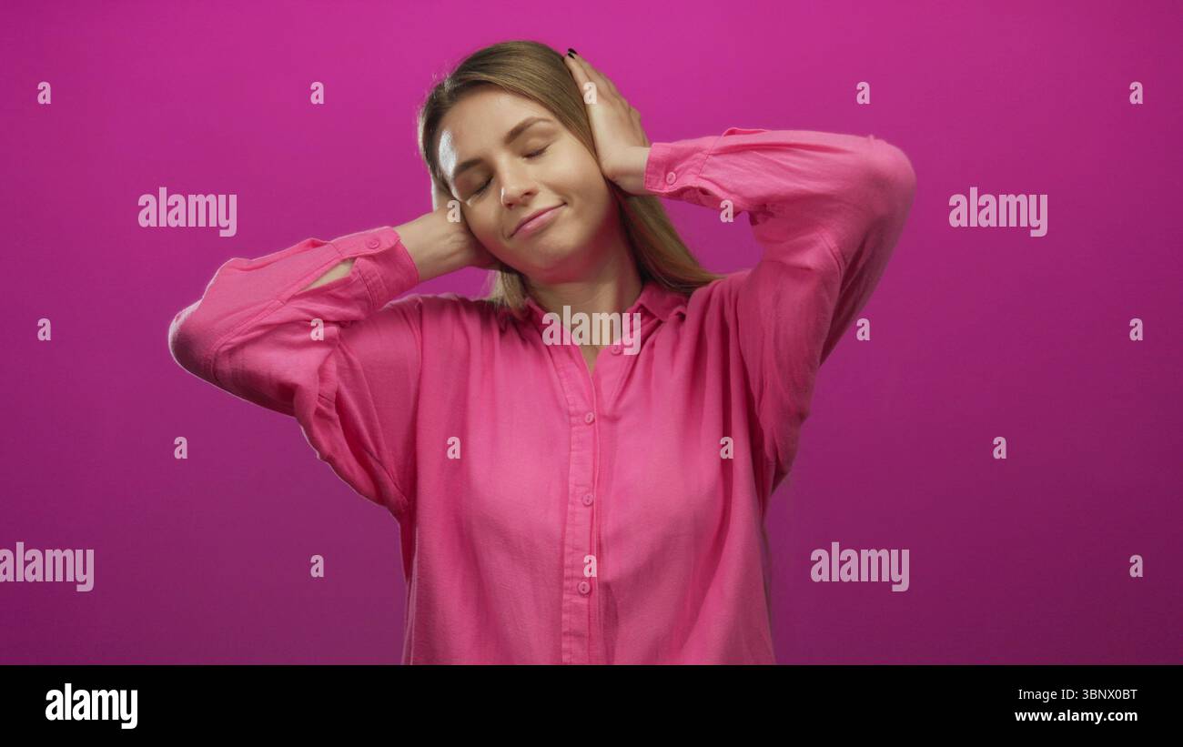 Young woman in pink shirt covers ears with hands, expressing frustration against a vivid pink background, conveying a sense of isolation and emotional Stock Photo