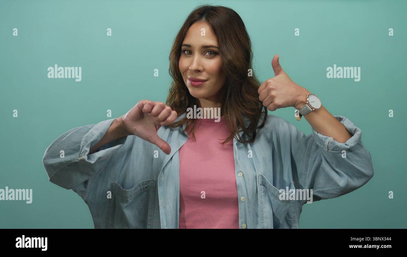Woman expressing mixed emotions with thumbs up and down against a vibrant green background, showcasing diverse communication and mood concept. Stock Photo