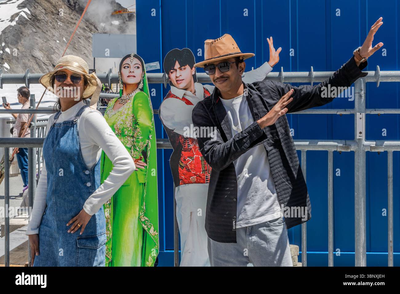 Between construction cranes and construction workers, Indian tourists pose in front of a cardboard cut-out of Bollywood actors Shah Rukh Khan and Kajol at the Titlis mountain station. The two actors are considered a famous screen couple who have starred together in numerous Bollywood films and shot romantic scenes in Switzerland. A couple of tourists pose on the Titlis in Engelberg, Obwalden, Switzerland, with cardboard cut-outs of Bollywood stars Shah Rukh Khan and Kajol. The man imitates Shah Rukh Khan's iconic pose. This place in the Swiss Alps is a favourite photo spot, especially for Indi Stock Photo