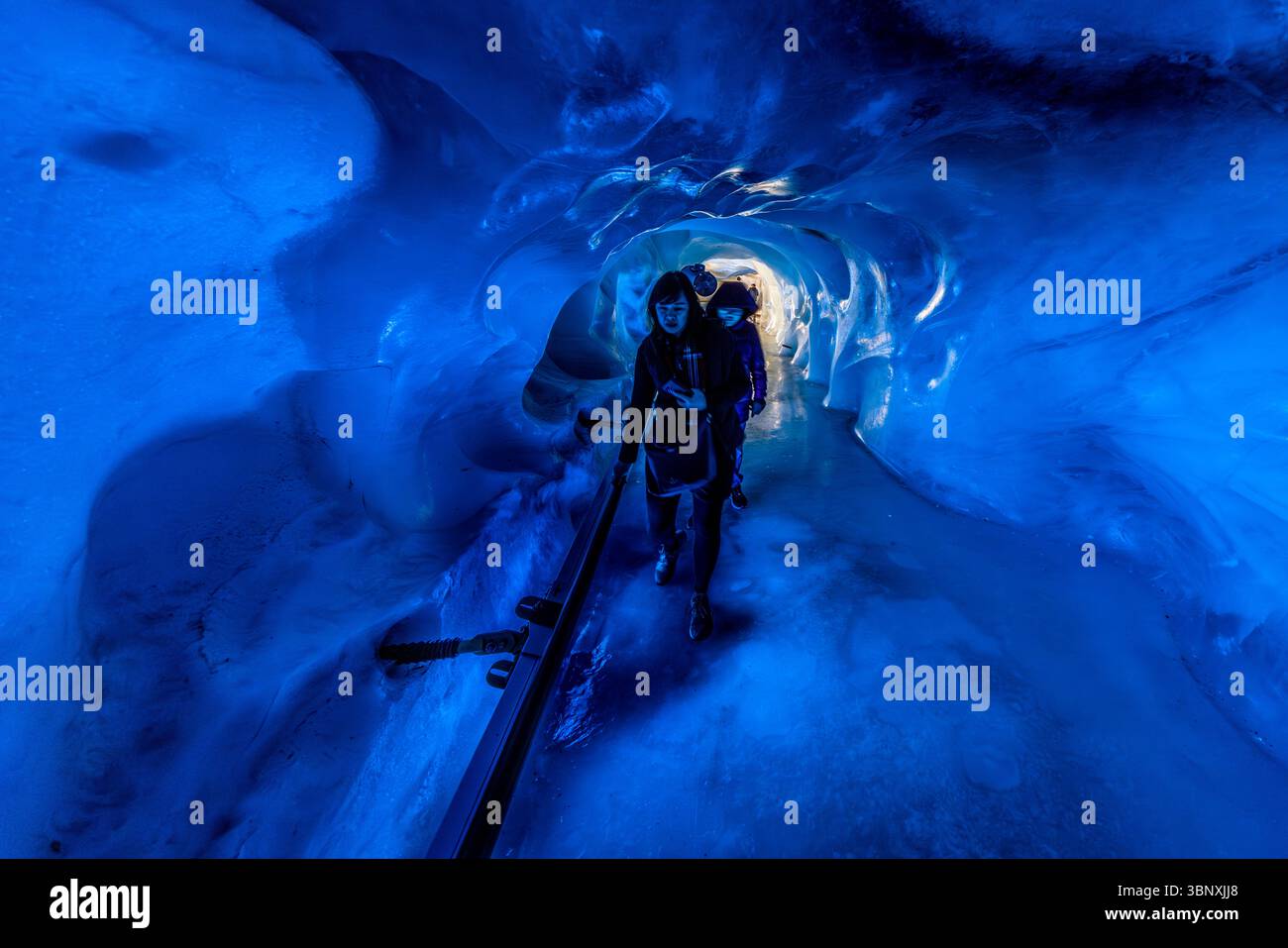The Titlis glacier grotto consists of an approximately 150-metre-long, artificially created passageway that leads around 20 metres below the surface of the glacier. The walls are covered with millions of glittering, ancient ice crystals, some of which are up to 5,000 years old. Two people explore the fascinating, blue-lit glacier grotto on the Titlis in Engelberg, Obwalden, Switzerland. They walk through the icy tunnel, which offers a unique experience in the Alpine landscape of Europe Stock Photo
