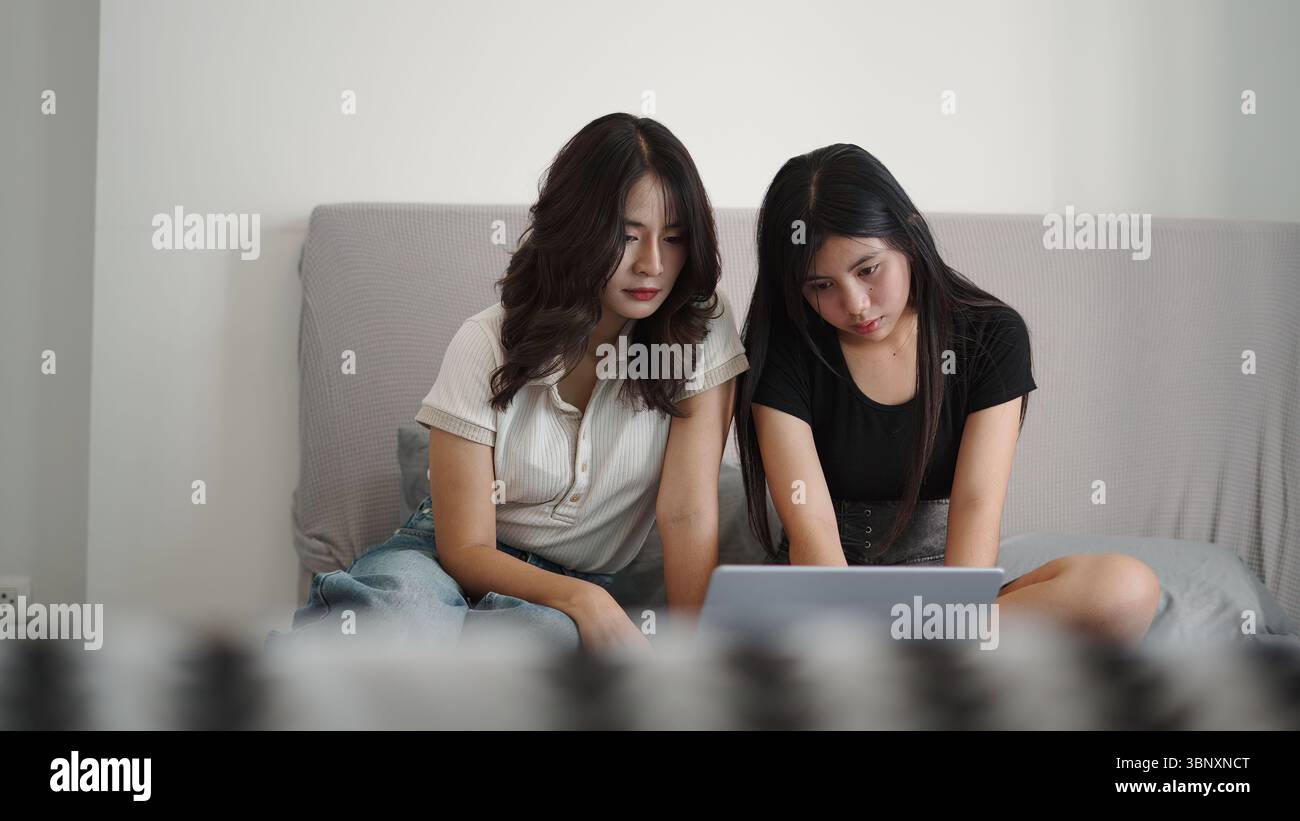 Two students are using a laptop, sitting on a bed, collaborating on a project Stock Photo