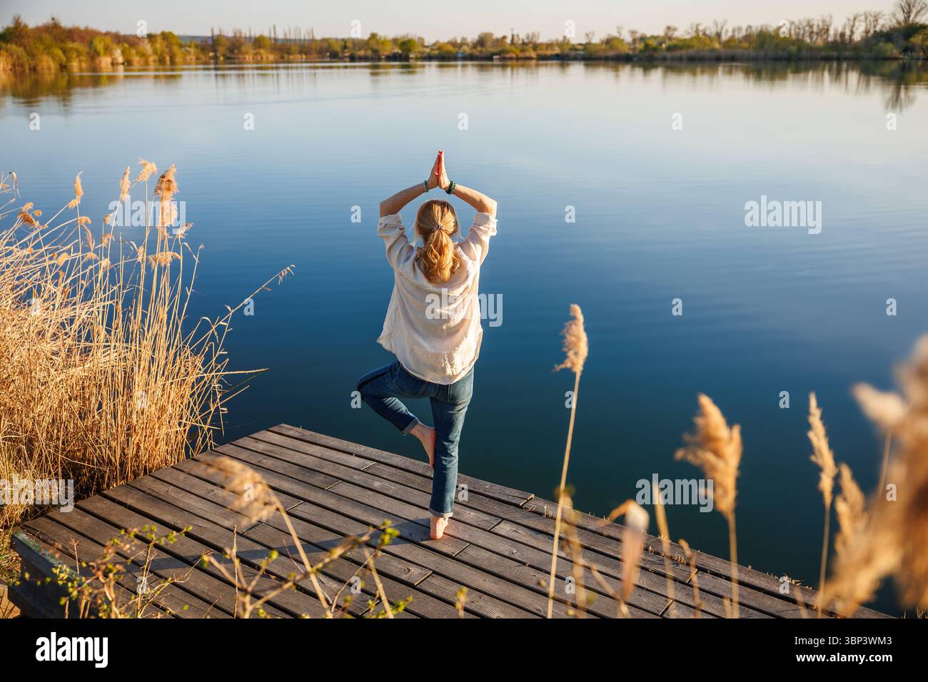 Woman practicing yoga exercise and tree pose on pier at lake. Fitness ...