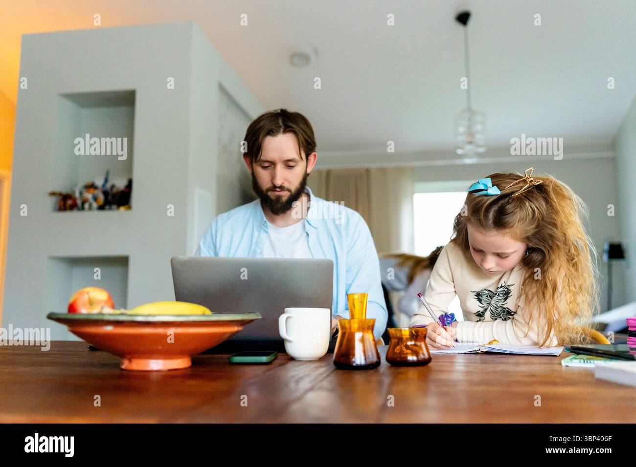 Father working on laptop while sitting next to daughter doing homework Stock Photo